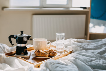 Fototapeta na wymiar Breakfast in bed: a wooden tray with croissants, glass of water and cup of coffee, on bed, backlit.