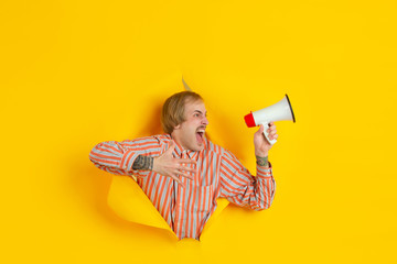 Shouting using speaker. Cheerful caucasian young man poses in torn yellow paper background, emotional expressive. Breaking on, breakthrought. Concept of human emotions, facial expression, sales, ad.