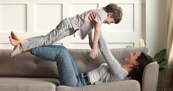 Full length smiling young mother lying on comfortable couch, lifting small kid son on straight hands indoors. Happy little child boy playing airplane with older sister on nanny in living room.