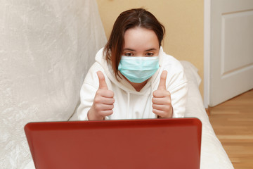 A young girl in a mask lies at home on a sofa with a computer and communicates via skype with...