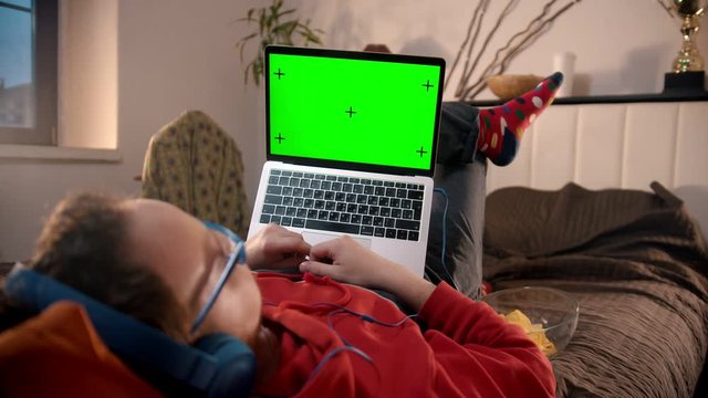 Young man lying on the bed with laptop and eating cheeps - green screen