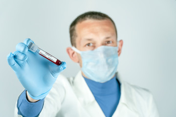 Male doctor in a medical gown, gloves and mask holds a test tube of blood with a positive result on COVID-19 close-up