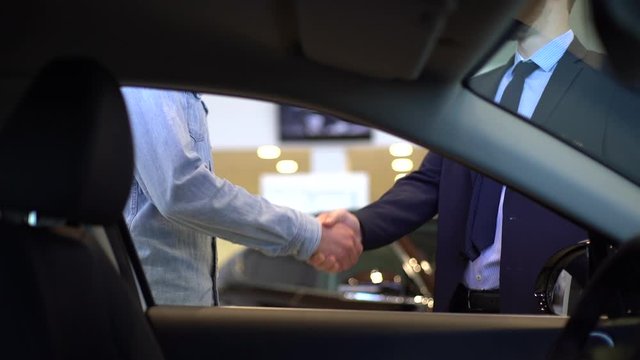 Close-up of unrecognizable salesman giving car keys to customer and shaking his hand in auto dealership, view from interior of car. Concept of buying new car at showroom. Shooting in slow motion.