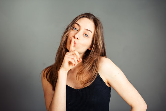 A girl in a black top with her hair is asking for peace and quiet, gesturing with a finger in front of her mouth, saying shhhh on a gray background. Without makeup, without retouching.
