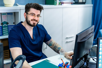Young medical worker sitting at desk. Bearded doctor in glasses smiling to camera in office.