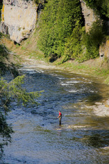 Young man fly fishing in a creek at the base of a large rock wall.