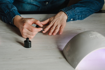 Woman doing manicure herself, draws, paints a brush, gel nail polish. Girl makes a manicure at home. Home life during quarantine, coronavirus pandemic, self-isolation. UV lamp with light. Top view.
