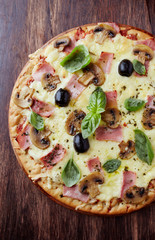 Tasty pizza with ham, champignon mushrooms, mozzarella cheese, black olives and fresh basil on wooden background. Top view.