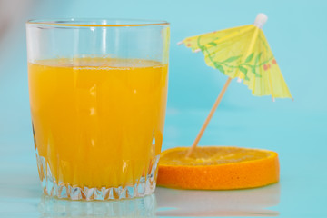 A glass with vitamin C-rich orange juice is next to a slice with drink umbrellas photographed against a heavenly background, a concept of natural quenching drink.