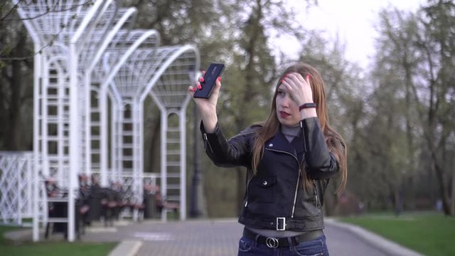 A red-haired Girl in a Black Leather Jacket Takes a Selfie with her Smartphone. Dependence on Gadgets. Psychological Disorders due to Other people's Opinions and Lack of Attention.