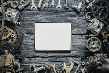 Blank screen digital tablet and old car spare parts on black wooden workbench flat lay background.