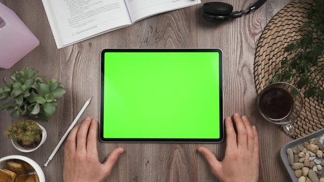 Horizontal orientation tab with green chromakey screen. Hand tap on center of the touchscreen then nervously waiting. Top view. Workplace table with many things. Daylight.