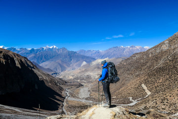 Fototapeta na wymiar Man hiking through dry path in Himalayan valley, located in Mustang region, Annapurna Circuit Trek in Nepal. He is having a short break, supporting himself on hiking sticks. Harsh landscape. Adventure