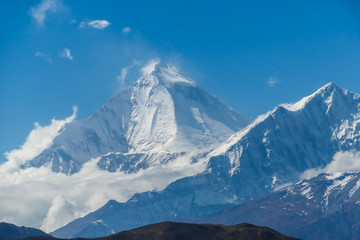Fototapeta na wymiar A distant view on snow capped Dhaulagiri I, seen from Mustang Valley, Annapurna Circuit Trek in Nepal. Wind blows the snow over the mountain peak. Barren and steep slopes. Harsh landscape.