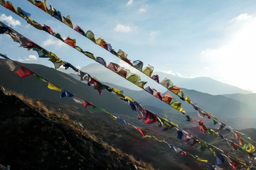 Prayer flags attached to the side of a mountain with a view on Himalayan chains shrouded in fog, seen from Muktinath, Annapurna Circuit Trek, Nepal. Sunbeams breaching through the peaks. Golden hour