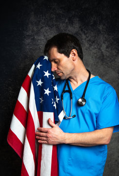 Healthcare concept - Doctor looking up for hope/cure, wearing blue hospital scrubs with a stethoscope, holding the American flag, and standing against a dark studio background.