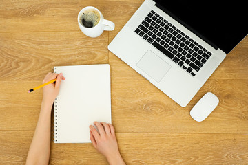 A composition on wooden background: a laptop, hands writing in a notebook, a cup of coffee and a mouse. 
