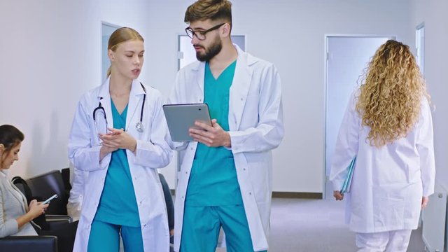 Modern hospital corridor two young doctors multiethnic female and man walking in front of the camera and discussing the future treatment for the patient while using the digital tablet concept of