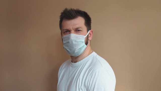 Handsome man in a white T-shirt puts on a medical protective mask and looks at the camera against a beige background. Portrait, the average plan. Natural light, soft focus.