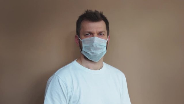Handsome man in a white T-shirt puts on a medical protective mask and looks at the camera against a beige background. Portrait, the average plan. Natural light, soft focus.