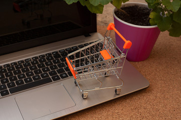 The concept of online shopping. On the table is a laptop, a mini cart of groceries and a potted flower.