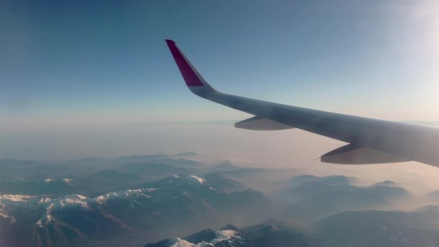 Wing of an airplane that flies over a beautiful view of the Swiss Alps. Fog and snow-capped mountain peaks, sunrise, sunlight