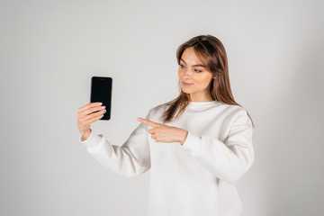 Fototapeta premium Cheerful cute woman pointing finger on smartphone screen isolated on a white background. Looking to the phone