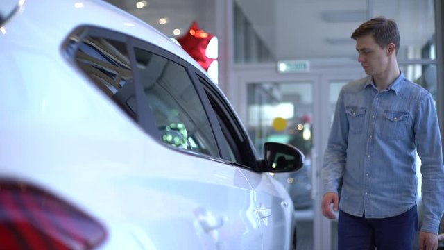 Handsome young man choosing new car to buy in auto dealership. Guy walking around the car dealership looking for new car. Concept of choosing and buying new car at showroom. Shooting in slow motion.