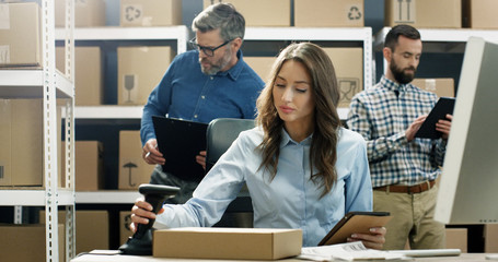 Woman working at computer in post delivery office and tapping on tablet device. Female postal worker scanning parcel with scanner and registering carton box. Mail co-workers on background.