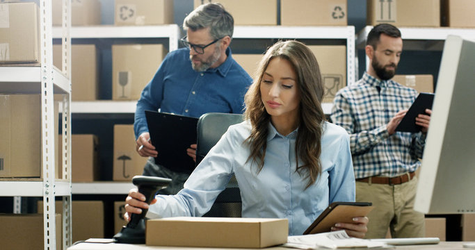 Woman working at computer in post delivery office and tapping on tablet device. Female postal worker scanning parcel with scanner and registering carton box. Mail co-workers on background.