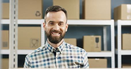 Close up of happy attractive young man smiling cheerfully to camera in postal office store among shelves with carton boxes. Portrait of male courier standing in mail storage.
