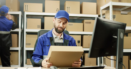 Portrait of delivery man in uniform working at computer in post office store with parcels. Postman...
