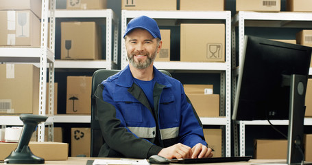Portrait of delivery man in uniform and cap sitting at table in postal office store and working at...