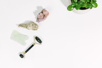 Obsidian jade roller, gua sha, sage smudging bundle, crystal, and succulent plant flat lay on white surface background