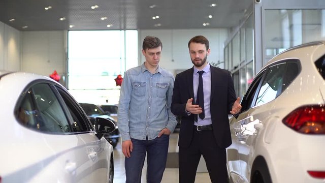 Confident car salesman talking and showing new car to handsome male customer in in auto dealership. Concept of choosing and buying new car at showroom. Shooting in slow motion.