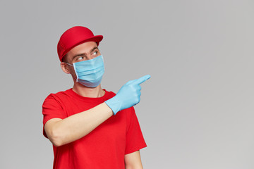 Courier in red uniform, protective mask and gloves points finger towards. Delivery service under quarantine. Space for text