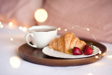 French baked croissant with ripe strawberry and black tea in white cup over glowing lights closeup. Summer season.