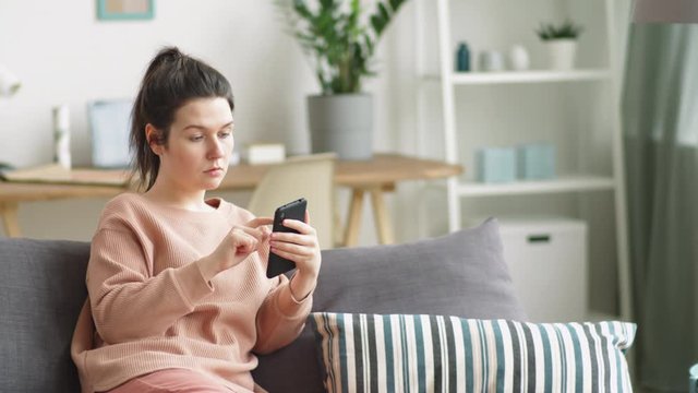Young Caucasian woman sitting on couch in living room and using smartphone while spending time at home