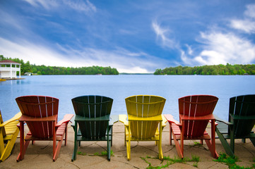 Colourful Adirondack chairs overlooking the blue water of a calm lake in Muskoka, Ontario Canada. A...