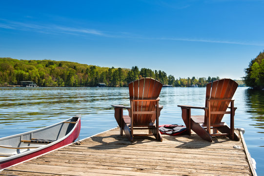 Two Adirondack chairs on a wooden dock on a lake in Muskoka, Ontario Canada. A red canoe is tied to the pier. Across the water cottages nestled between green trees are visible.