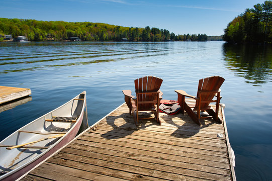 Two Adirondack chairs on a wooden dock facing the blue water of a lake in Muskoka, Ontario Canada. A red canoe is tied to the pier. Across the water cottages nestled between green trees are visible.