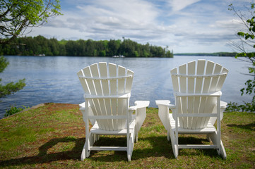 Fototapeta premium Two white Muskoka chairs overlooking at a lake in Ontario Canada. Across the calm water there's a cottage nestled between green trees.