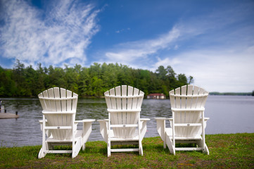 Three white Muskoka chairs overlooking at a lake in Ontario Canada. Across the calm water there's a cottage nestled between green trees.