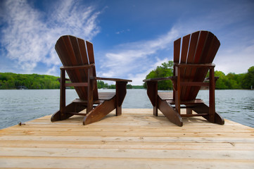 Two Adirondack chairs on a wooden dock overlooking a calm lake. 
