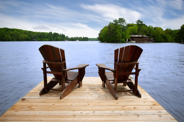 Two Adirondack chairs on a wooden dock overlooking a calm lake. A brown cottage nestled between...