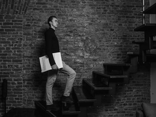Full length portrait of a handsome stylish man standing on stairs with newspaper and posing in loft interior. Monochrome