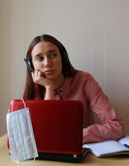 A girl with headphones dreamily sits at a table with a computer and copybooks. Distance education. Schools and universities are on quarantine due to the pandemic of coronavirus Covid-19.