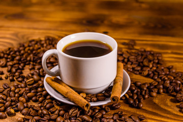 Cup of hot coffee, cinnamon sticks and scattered coffee beans on wooden table