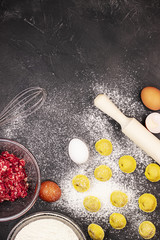 Dark marble background strewn with white flour with eggs, dough, minced meat and a rolling pin, top view. Concept of cooking dumplings