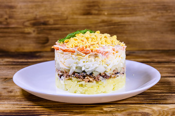 Plate with traditional russian salad Mimosa on a wooden table. Layered salad with potatoes, sardine, cheese, carrot, eggs and mayonnaise
